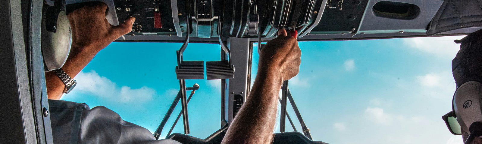 Scene from the cockpit in a plane; two pilots partly seen from the back, one adjusting flight instruments.