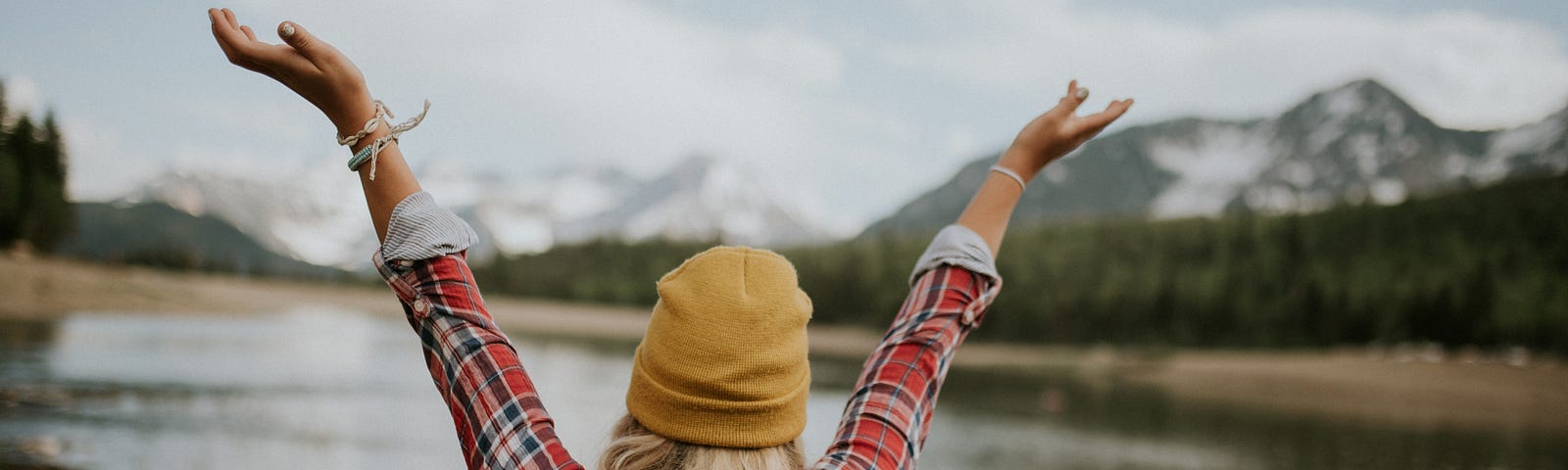 A blonde woman facing a body of water and mountains with her arms raised