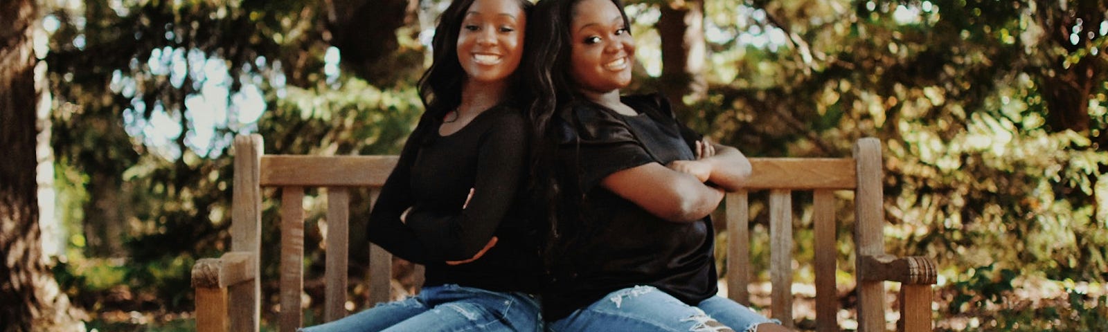 Young adult twins wearing jeans and black shirts sit with their backs together on a wooden bench. Trees are in the background.