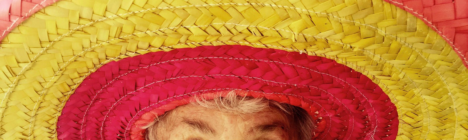 An older woman with a funny look on her face looks into the camera lens from under a giant red and yellow sombrero.