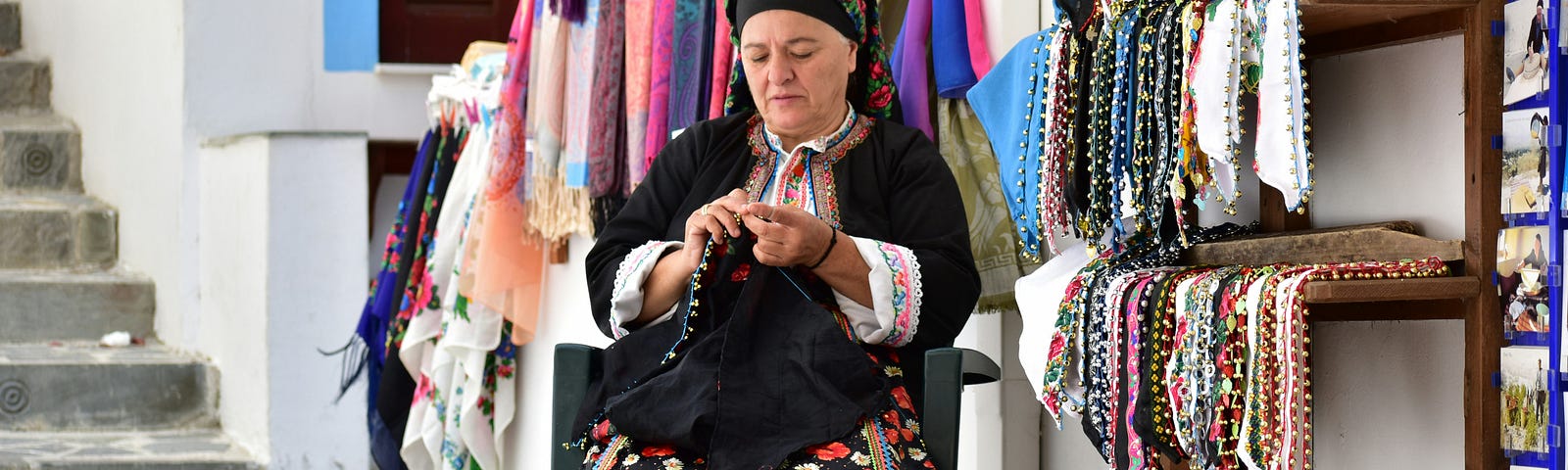 An Eastern-European woman in colorful garb sits outside a store and sews