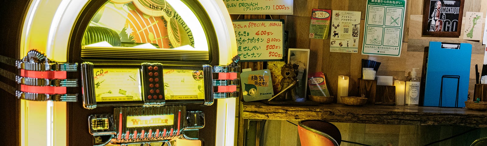 A cluttered picture of a cafe. A juke box is featured prominently on the left side of the picture. There are menus written on dry erase boards on the wall, a counter full of items and a chair.