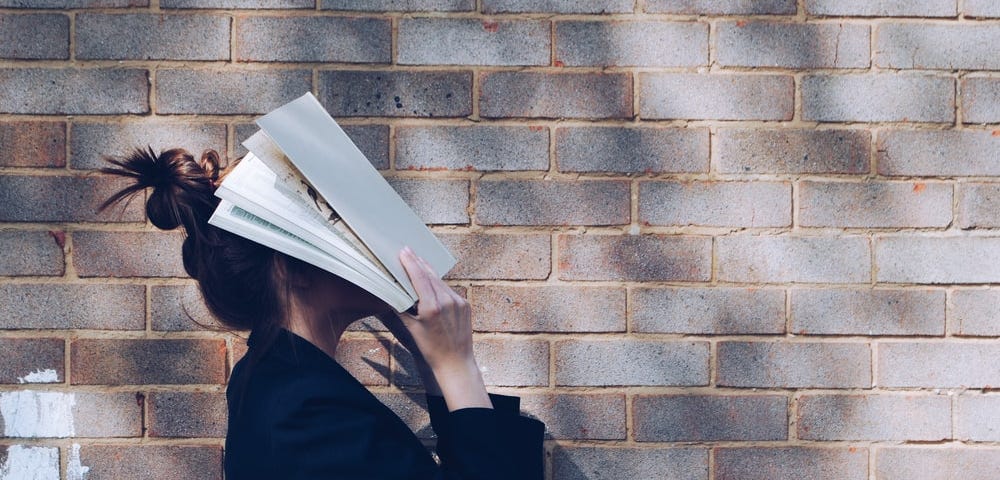 School Girl with Book in front of natural rustic red brick background holding book up to her face.