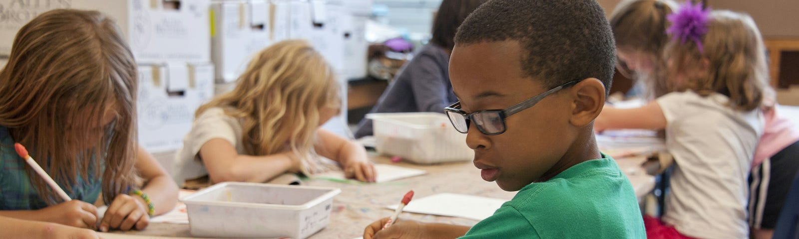 Children in school at a table writing and drawing with a black-skinned boy in a green T-shirt in the foreground and brown and blonde-haired girls in the background.