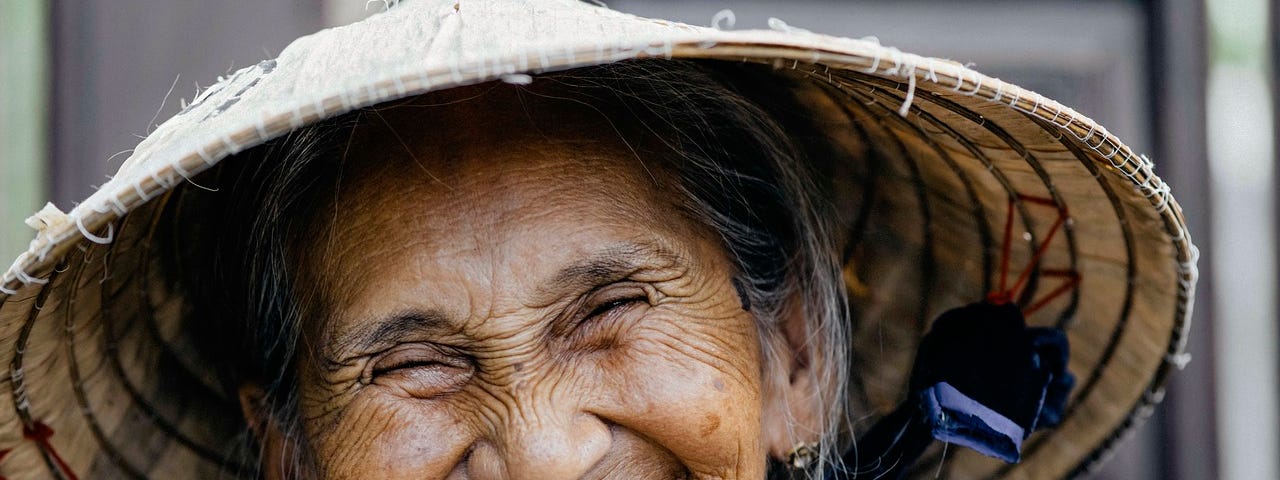 Elderly dark-skinned woman with a conical Asian hat, smiling broadly, causing her eyes to squint and nose to wrinkle. This woman exudes joy in her smile.