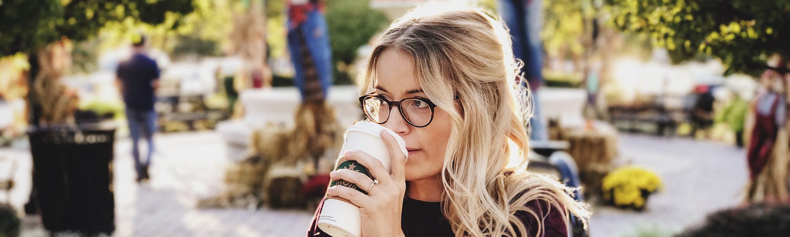 Blonde woman with eyeglasses sipping coffee while walking along a park