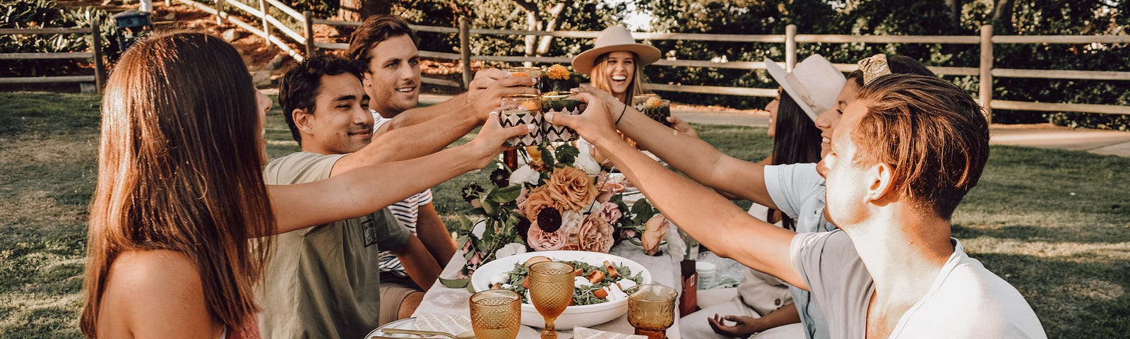 A group of seven people toasting at a picnic table outdoors. There is a whole spread of fruit, crackers, and cheese on the table, along with flowers for decoration. The photo has a golden hue, like it was taken during sunset.