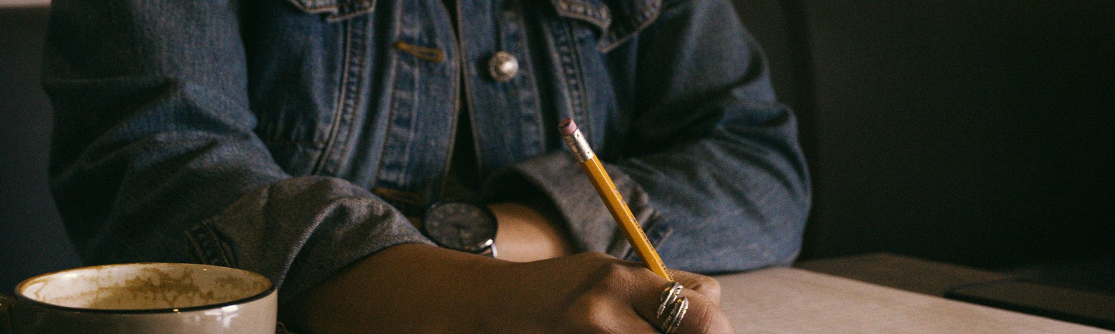 Brown skinned black woman in denim jacket writing in notebook with a pencil. A cup of coffee and two books rest on the table.