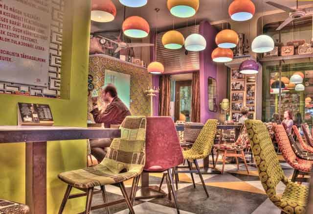 Hipster cafe with man seated in one of many colorful, pattered ulpulstered chairs and big round light fixtures above.