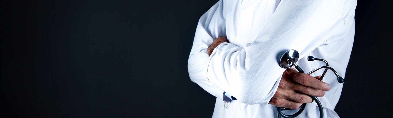 A black doctor in white coat stands with arms crossed on the right half of the photograph, his stethoscope curled in his right hand. We see him from the waist level up to mid chest. Black background.