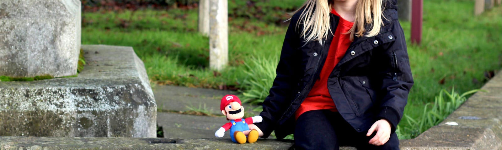 A sad looking young girl with blond hair is sitting on a stone bench in a cemetery