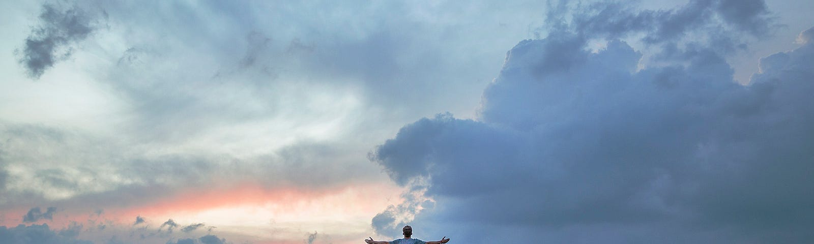 A person stands, arms outstretched, atop a rocky outcrop, facing the landscape as dusk approaches