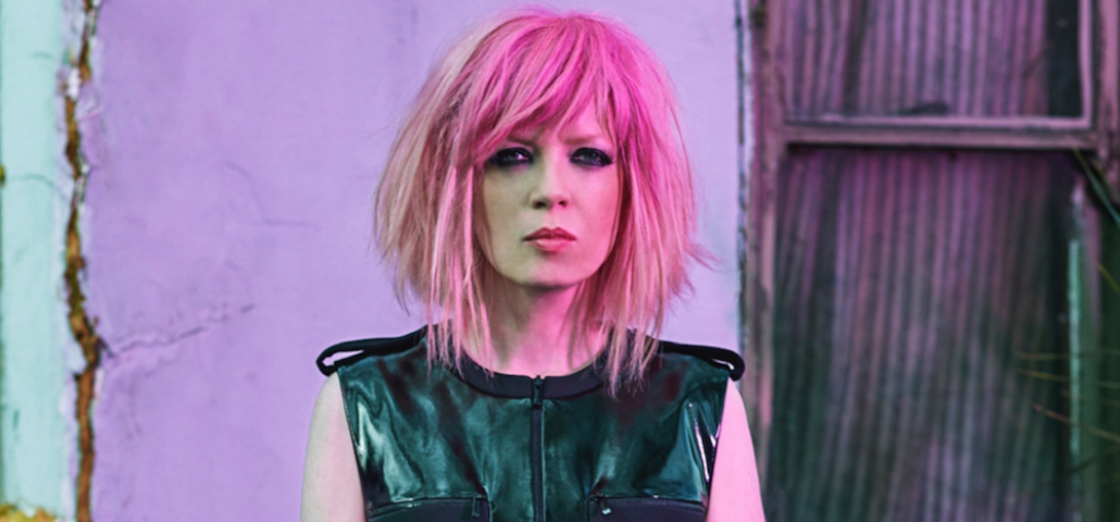 A photograph of Shirley Manson, lead singer of American band Garbage. She is standing in front of a backdrop of concrete and corrugated steel. She has pale skin, and neck-length pink hair. She is wearing a black sleeveless top with patches of different material and a front zipper. Her facial expression is neutral and unsmiling. She is looking directly at the camera.