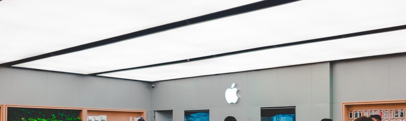 The image depicts the interior of an Apple Store. You can see the iconic minimalist design with wooden tables displaying products. There are several customers and staff members inside, some are looking at products, and others are conversing. Above, large, bright ceiling panels illuminate the space, and the Apple logo is visible in the background, glowing on a wall panel. The setting is typical of Apple’s retail locations, known for their open layouts and hands-on product displays.