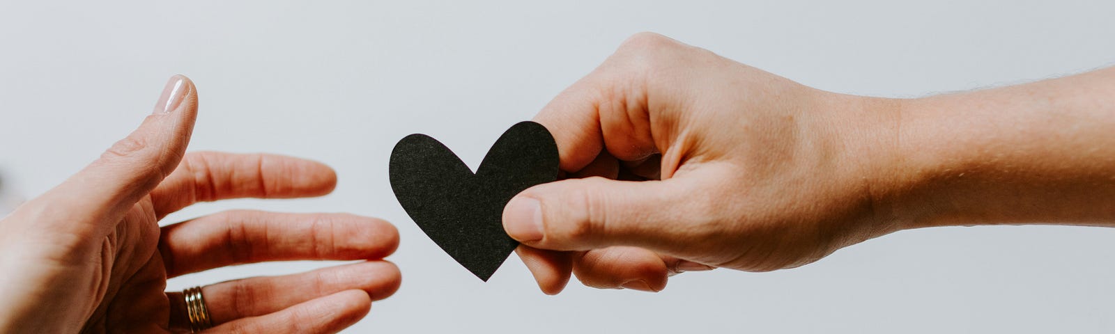 Showing only their hands, one person offers a paper cut-out of a heart to another person, who reaches to receive it.
