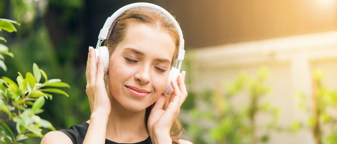 A white woman with red hair is standing outside by tall plants at sunset. She’s listening to music using headphones and smiling.