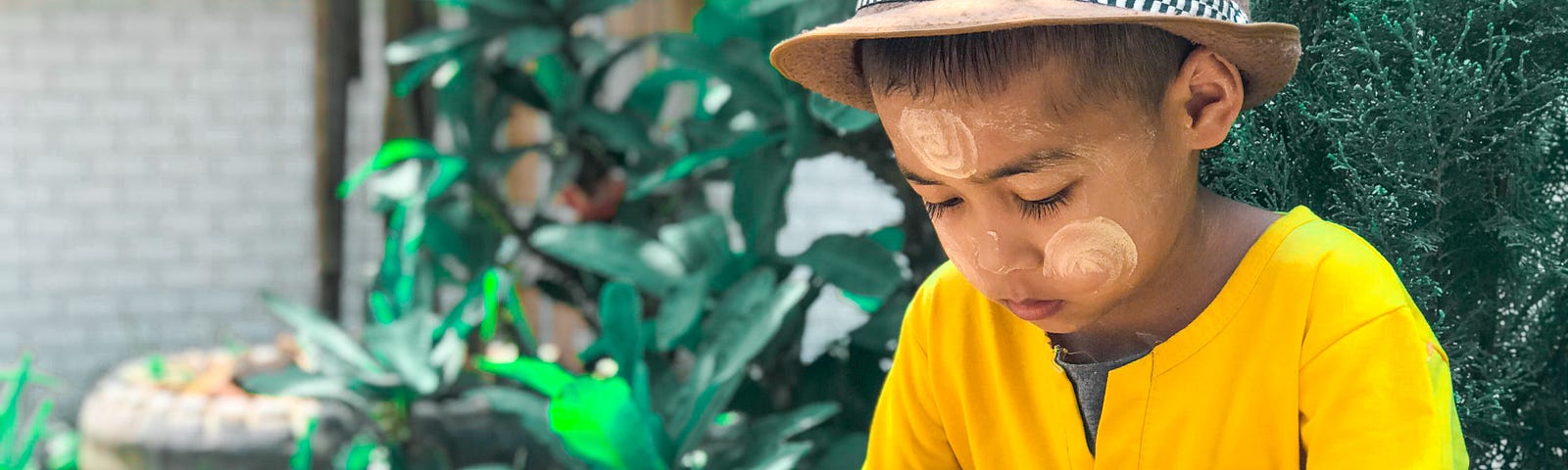 A little boy sits in the garden looking at a phone unsupervised. He wears a brown bowler hat, yellow t-shirt and shorts.