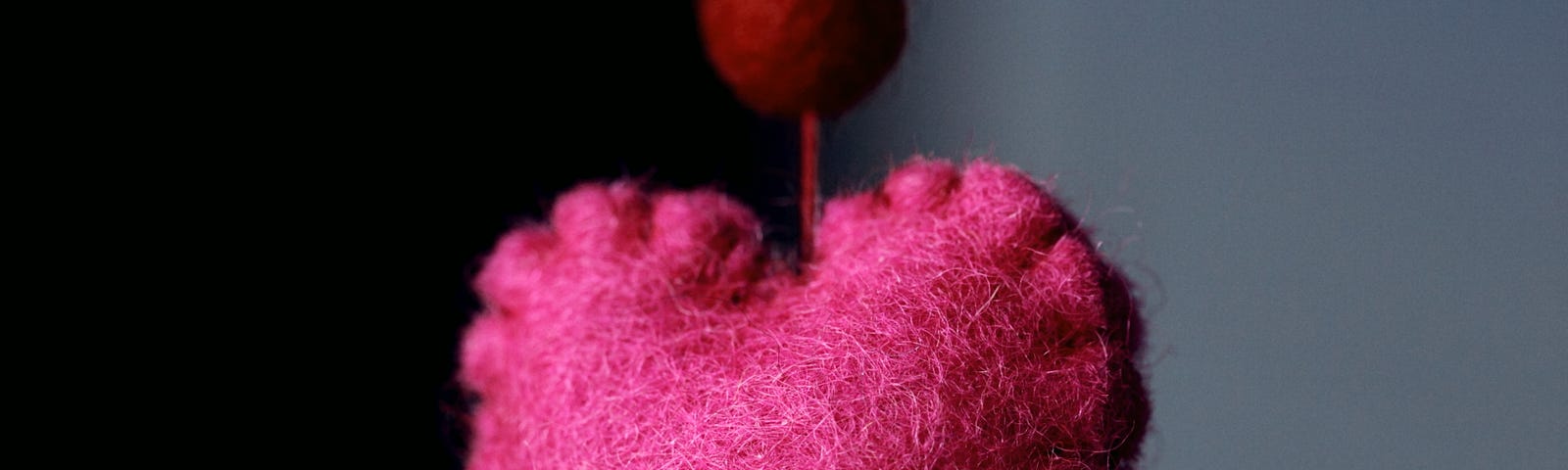 A close-up of a crafted pink heart on a string against a backdrop that is half black and half gray.