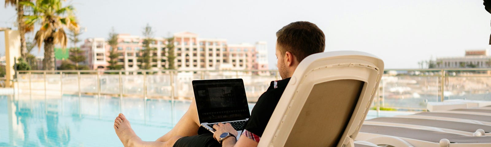 A digital nomad man sitting before a large swimming pool, working on his laptop