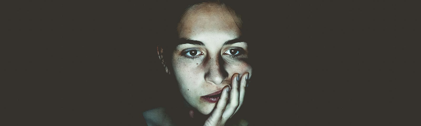 woman sitting in the dark, head resting in her hand, staring boredly at a screen.