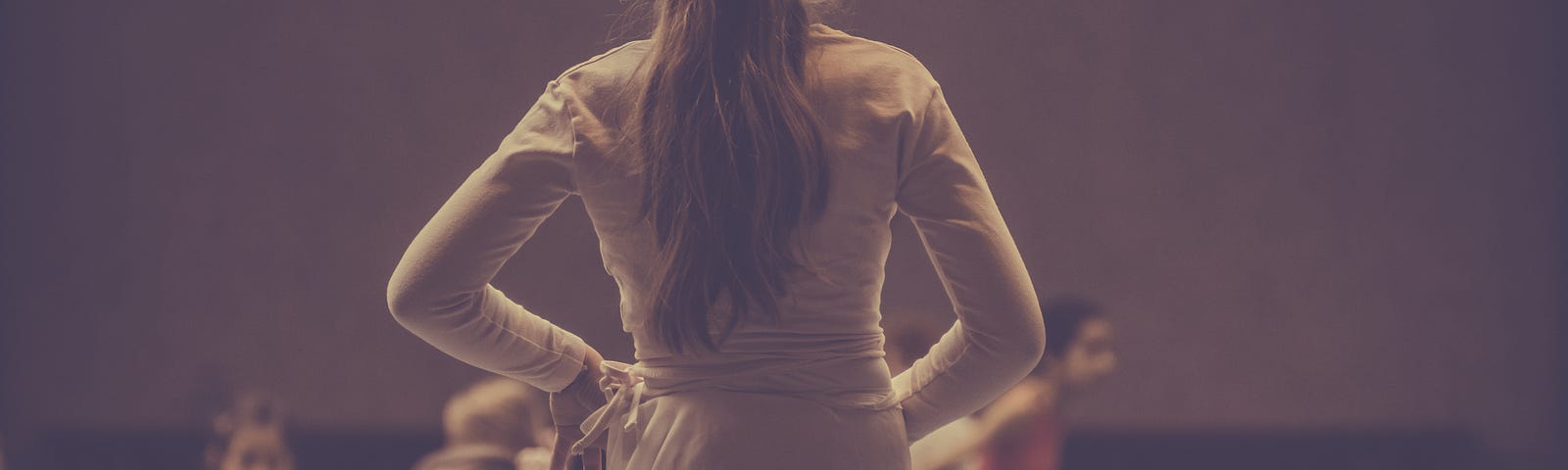 A photo of the back of a young girl singled out standing in a plié, practicing ballet, in front of other girls sitting on the mat.