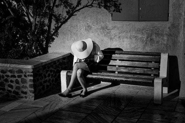 A woman wearing a hat is sitting alone on a bench alongside a wall. She has her head down and is obviously depressed.