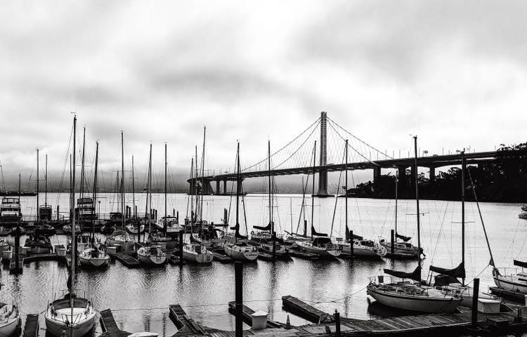 A black-and-white photograph of a marina on a cloudy day. Multiple docked boats are in the foreground and the San Francisco Oakland Bay Bridge is in the background.