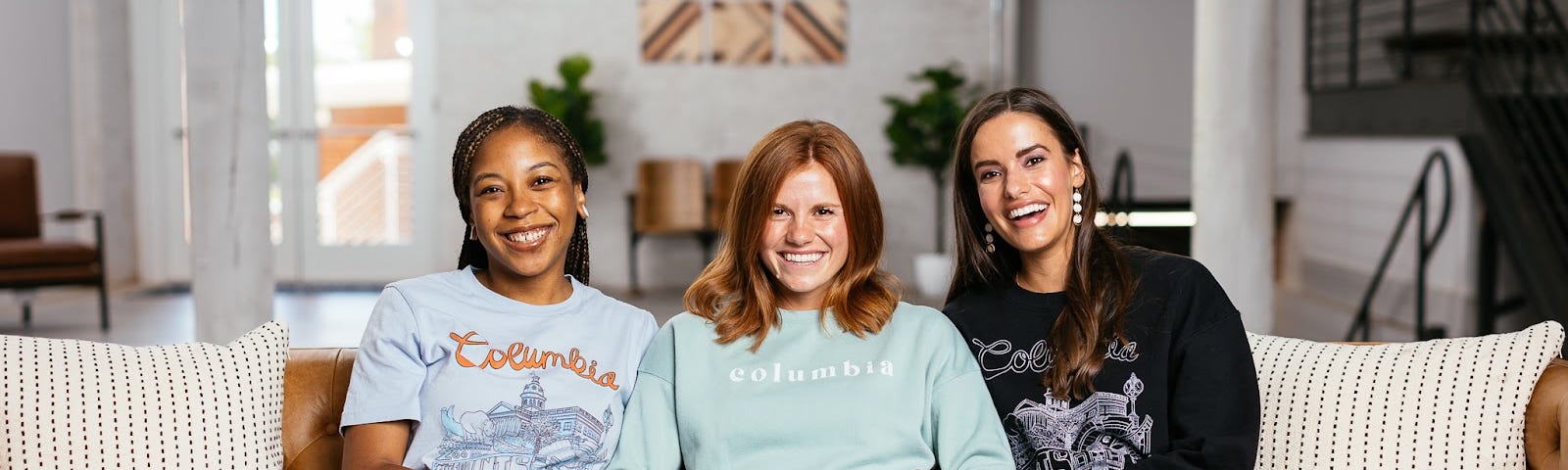Three women, one Black and two white, sit on a leather couch in a bright, airy loft-style space. They model Six & Main long- and short-sleeved shirts with Columbia, South Carolina-specific designs on them, including the city name and landmarks.