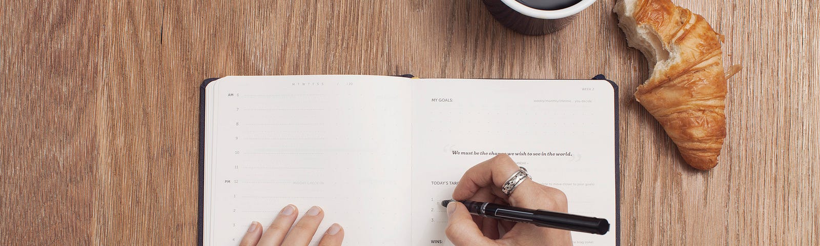 Hands of a woman writing on a notebook, a cup of coffee, and a croissant