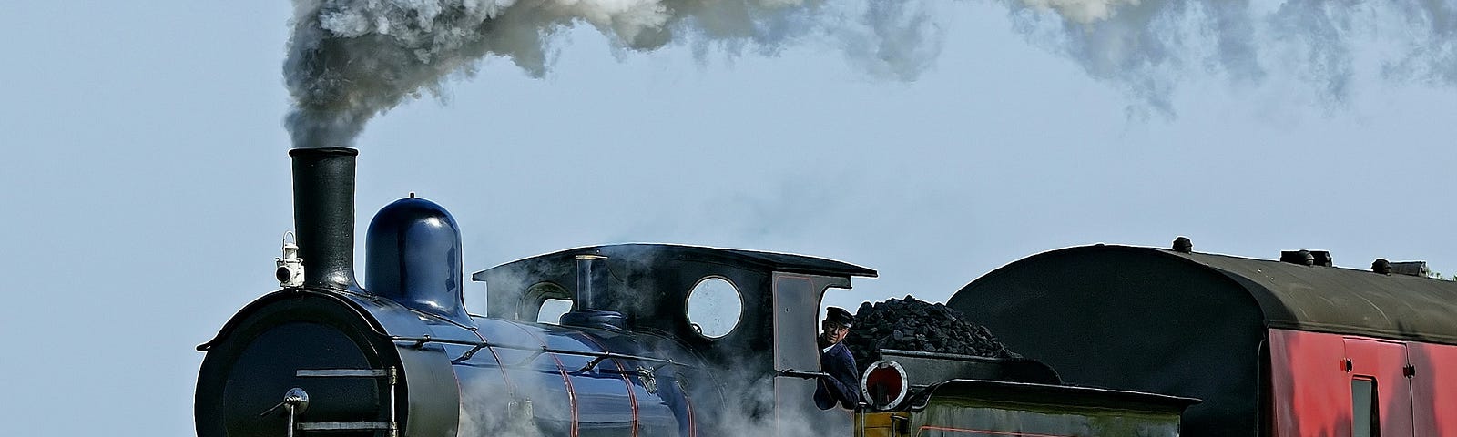 Image of a black steam train locomotive, blowing out smoke and you can see a part of the red carriage behind it.
