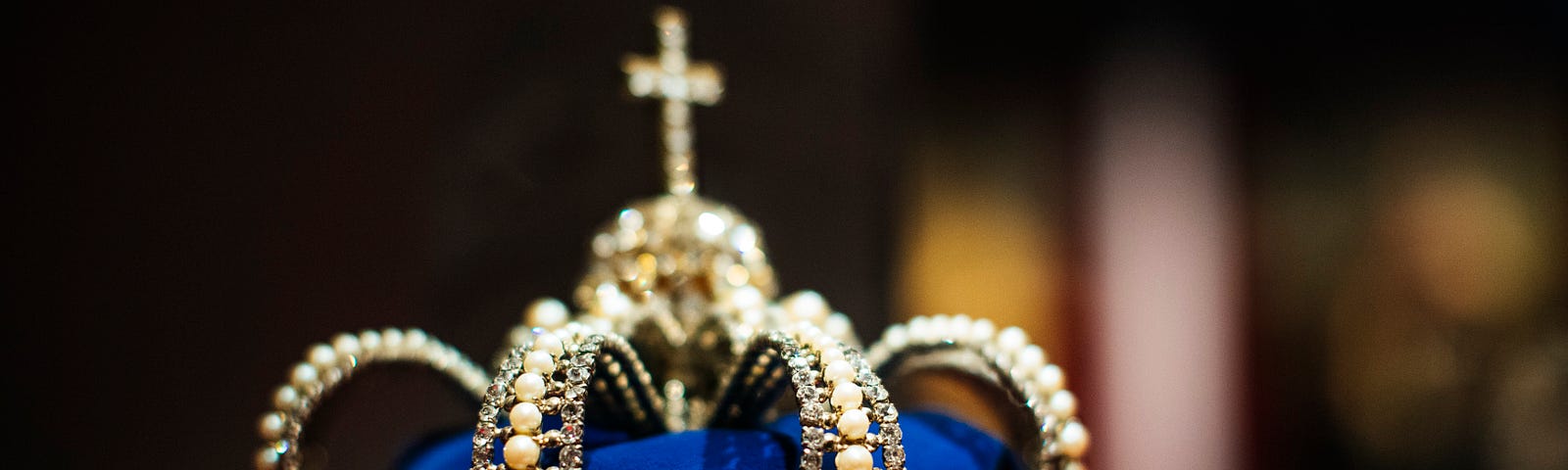 A crown made of pearls and diamonds over deep blue fabric with a crucifix on top sitting on a red pillow