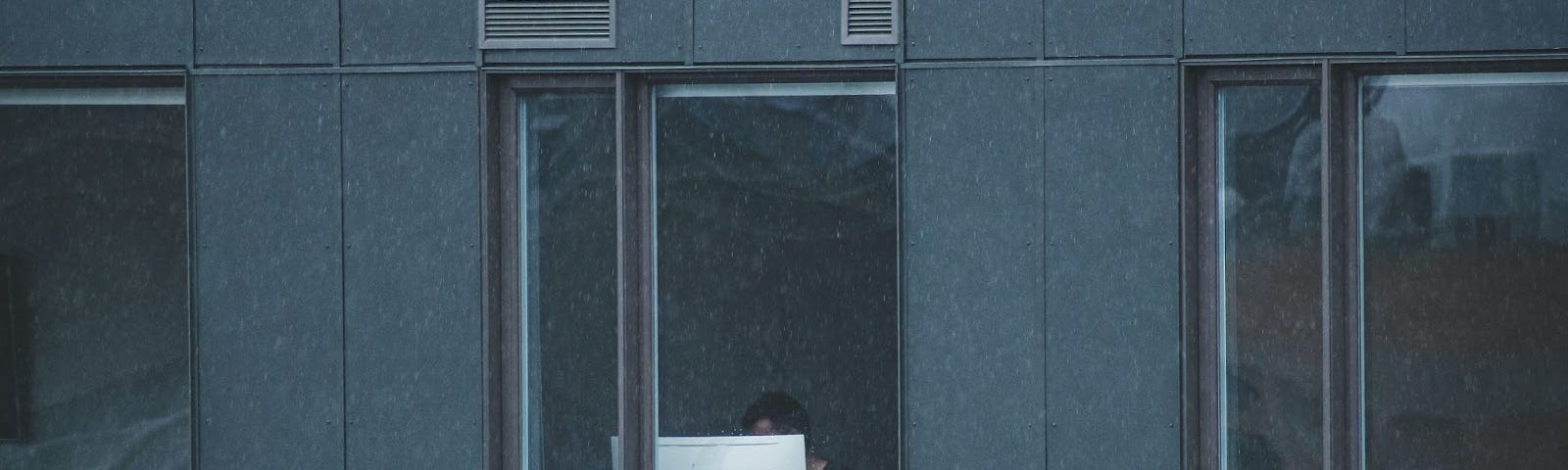 vista de fora de um homem trabalhando no computador em frente à janela. O prédio é cinza e o monitor se destaca por ser branco. | portainer ubuntu contêineres docker