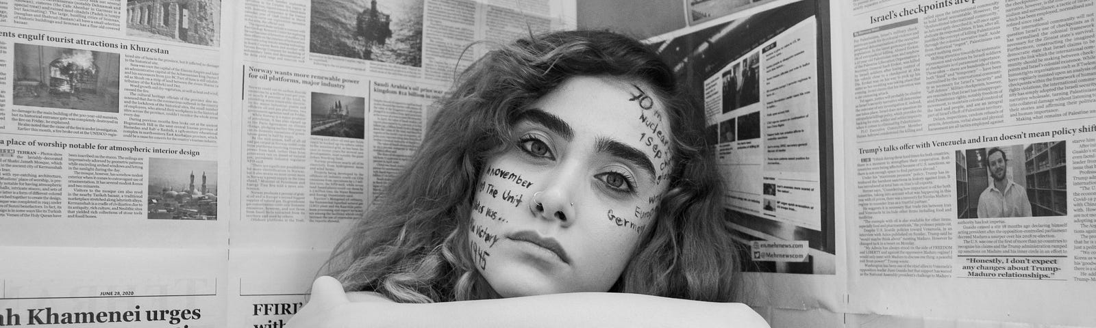 black and white scene of a young woman with writing on her face, hugging her knees. there are newspapers on the walls on either side of her