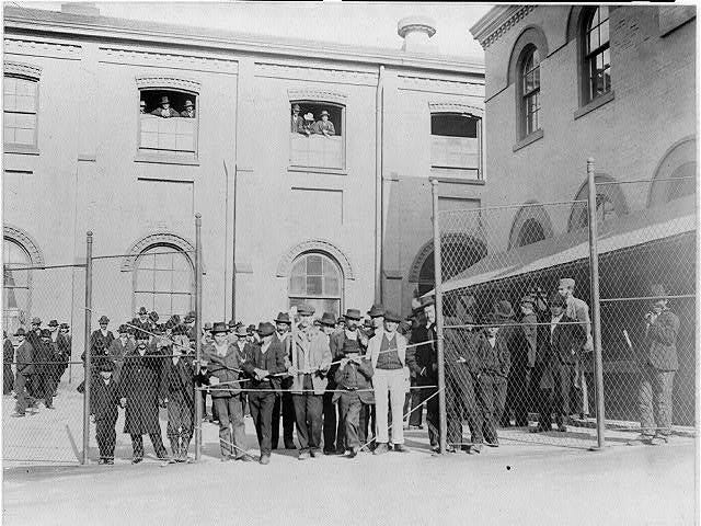 A group of people stand behind a gate and a fence in front of a building. The image is black and white, and most of the people are men. They are wearing clothes from the turn of the twentieth century.