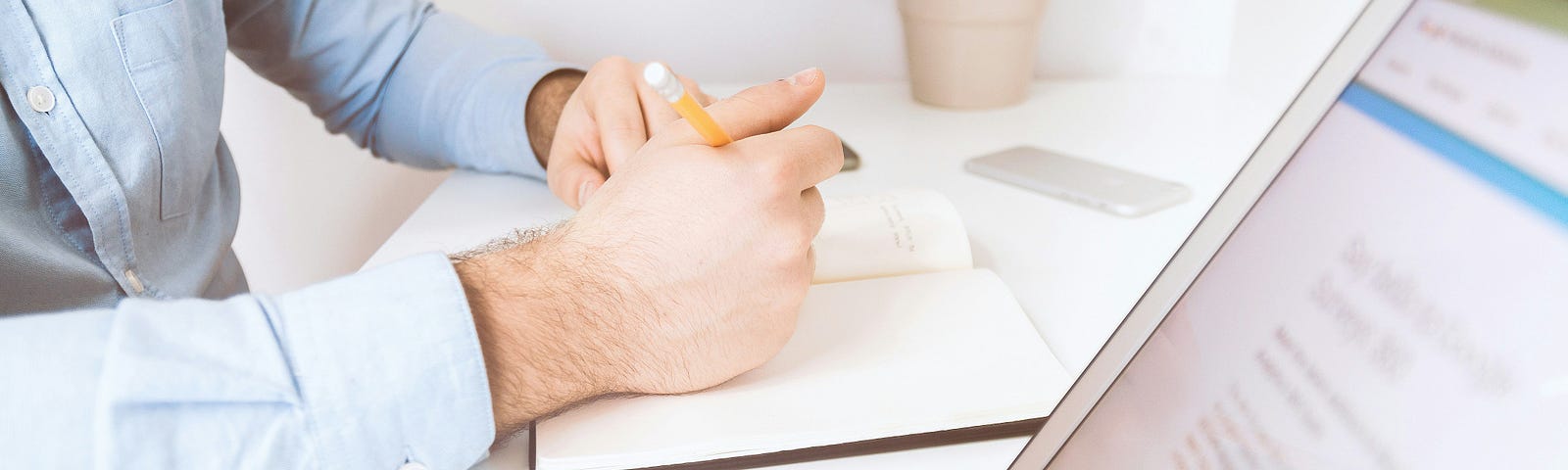 Man at a desk writing in journal with open laptop showing