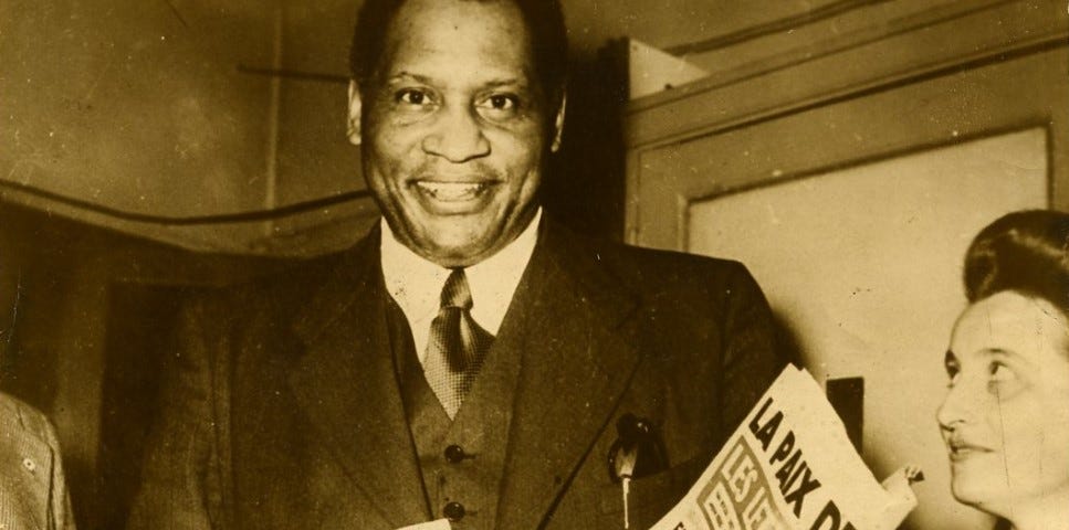 Smiling very tall man wearing a suit and carrying a newspaper. To his right and half cropped from the photograph is a woman smiling and looking upwards towards him