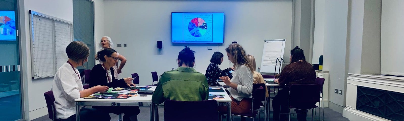 Seven participants sitting around white tables with archive materials and contributing in the Shaping Narratives Workshop at Wellcome Collection.