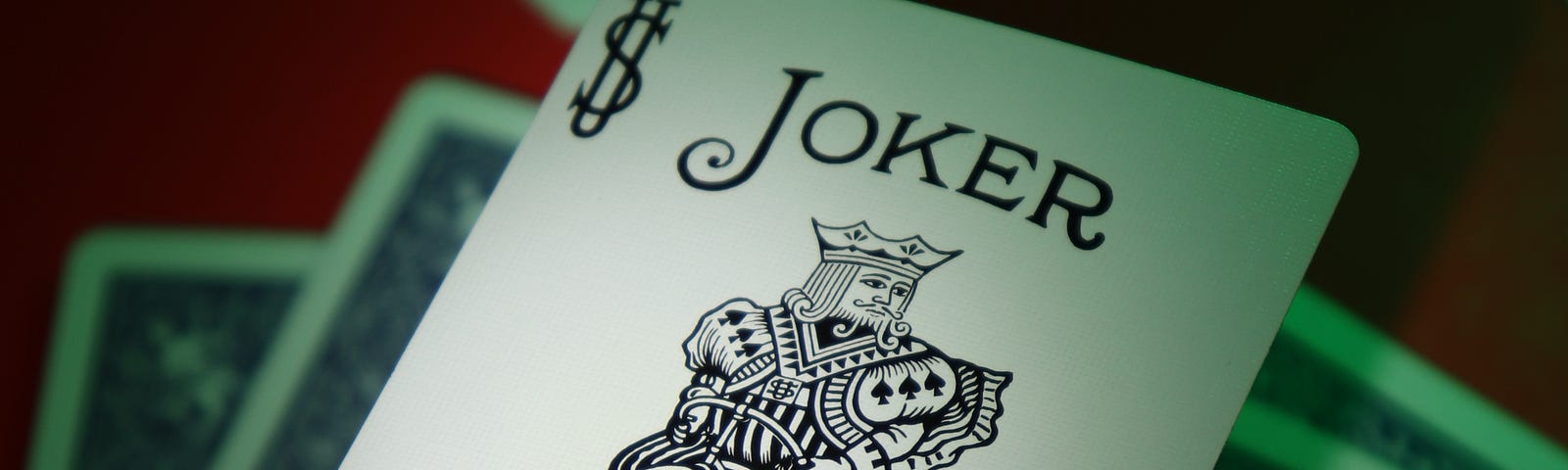 Joker card, atop several face-down cards, black-on-white image featuring line-drawing of figure on bicycle; red background.