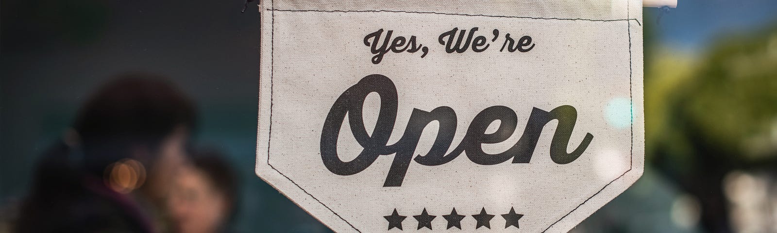 Image of a small banner on a glass door reading “Yes, We’re Open”