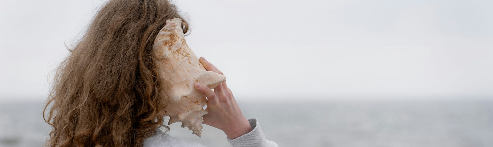 Woman listening to the sound of the sea in a conch shell, while she stands on a beach.