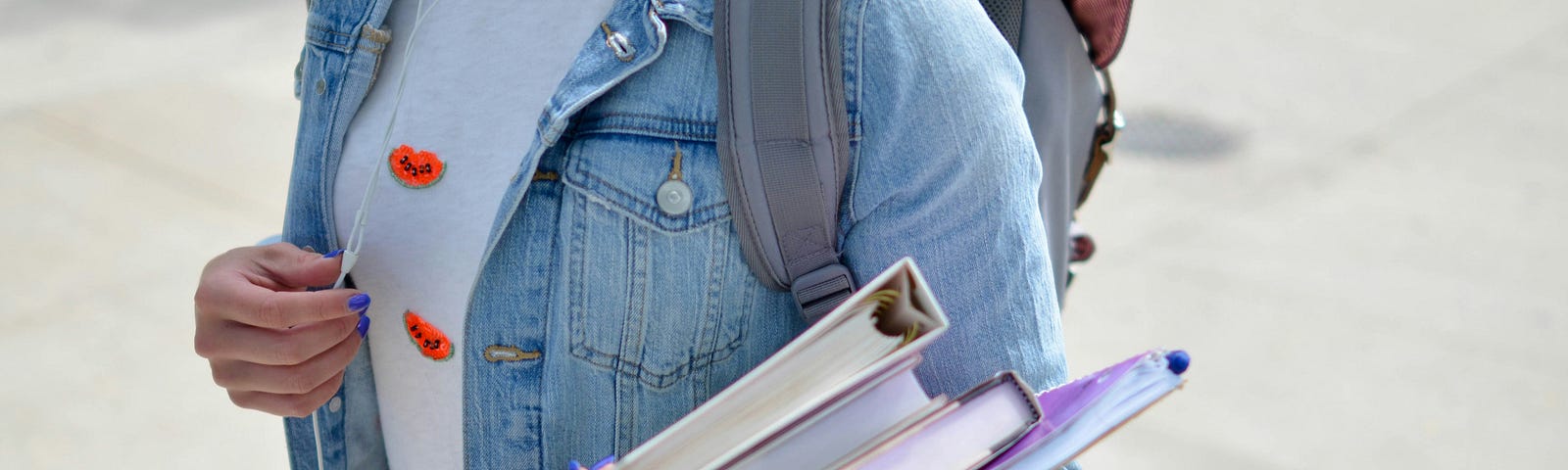 Student on the sidewalk with books on her arm and headphones in her ears.