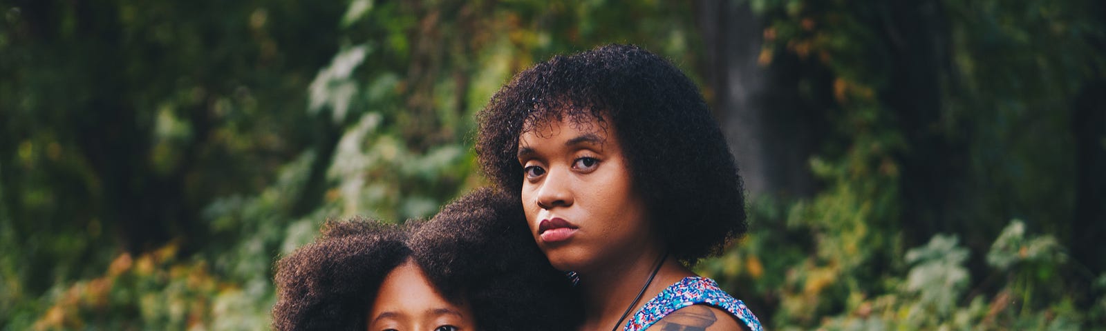 A Black mother and daughter are sitting on the ground in a forest. The daughter is leaning backwards on her mother. They both have serious looks on their faces.