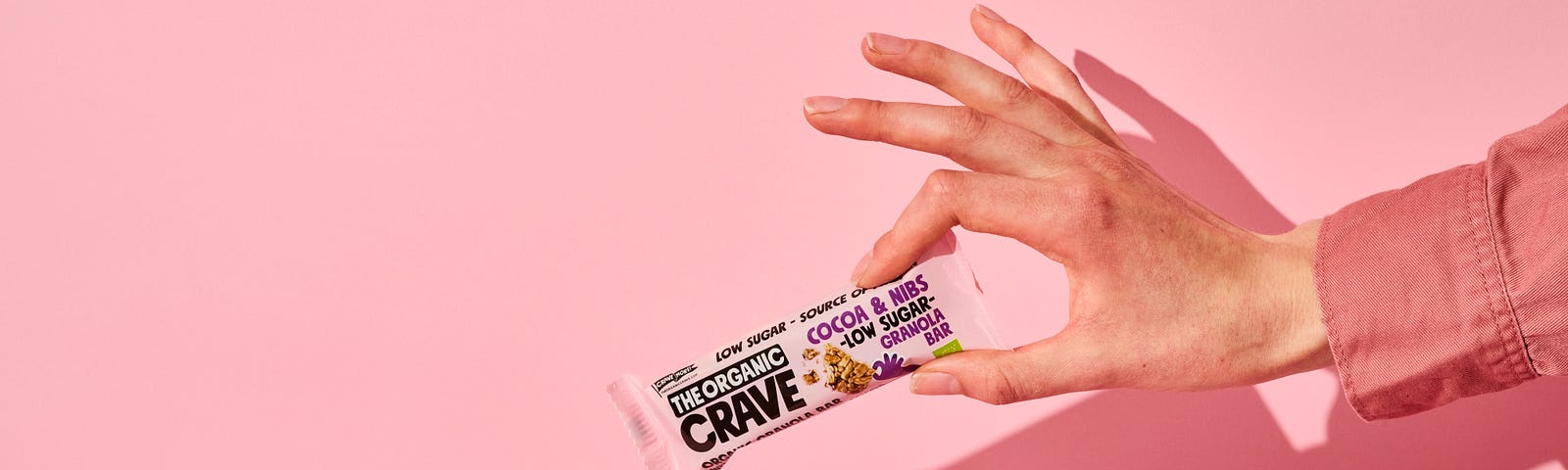 A hand emerges from the right to hold a protein bar (unopened) with his/her thumb and index finger. Pink background.