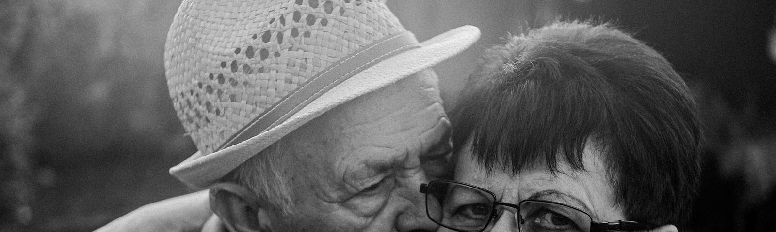 A black and white photo of an elderly man and woman. He is wearing a white hat and she’s wearing glasses.