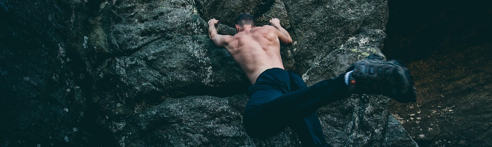 A topless muscular man in dark track-suit bottoms hangs from a rock with his bare hands, his back towards the camera.