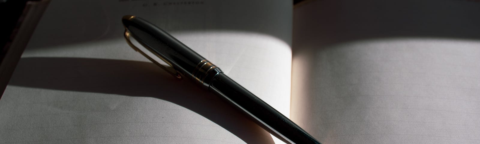 A pen lying across a blank notebook and casting a shadow.