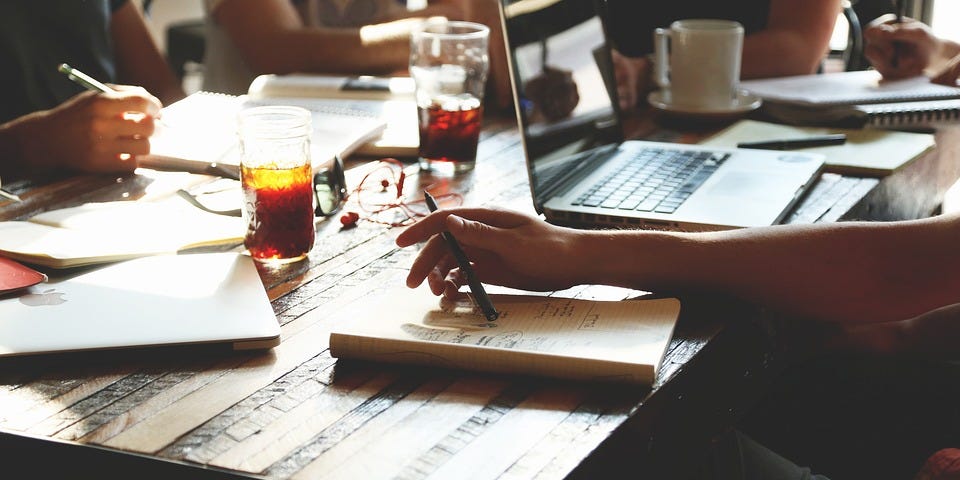 A table where four people sit and work on a project. On the table, we find a laptop and notebooks the participants are using.