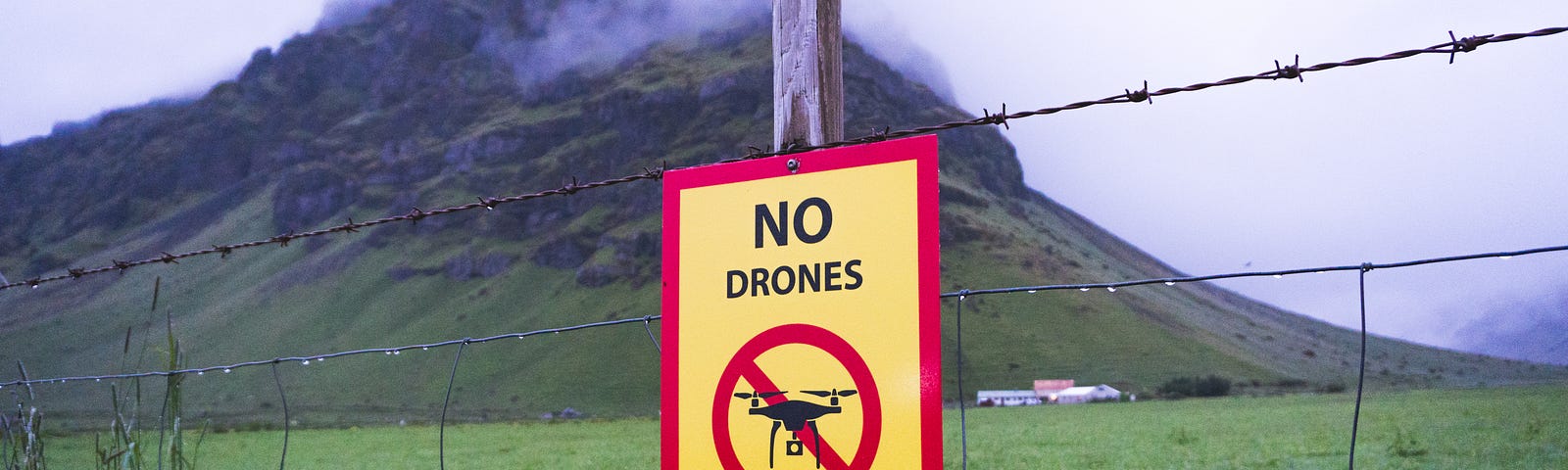 A barbed wire fence in front of a steep hill with some cloud cover at the top. The sign on the fence says “NO Drones.” There are some low buildings at the foot of the hill.