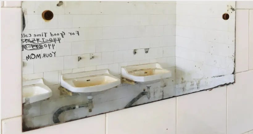 Old, corroded, public bathroom mirror reflecting an image of dirty sinks missing fixtures and the aged, white concrete block wall with lines of graffiti written in black that are reversed in the mirror reflection.