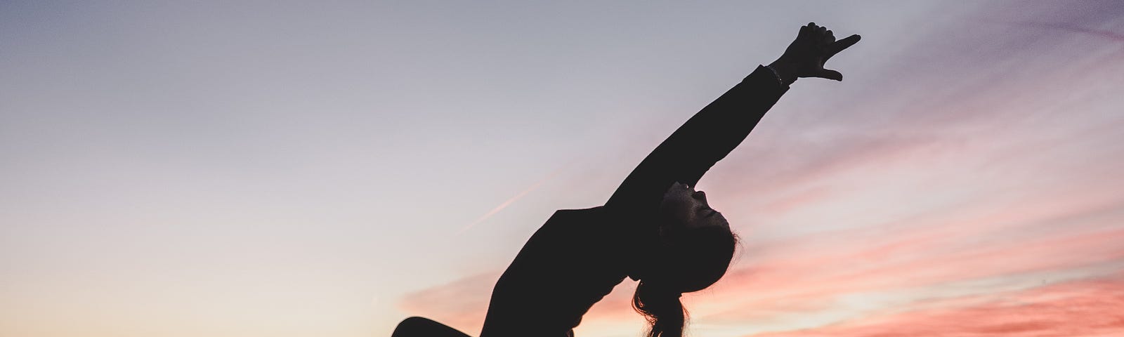 Woman facing to the left of the screen practices yoga. Her right knee is forward, and her left leg extended behind her on the ground as she bends backward, arms overhead. Sunset in the background.